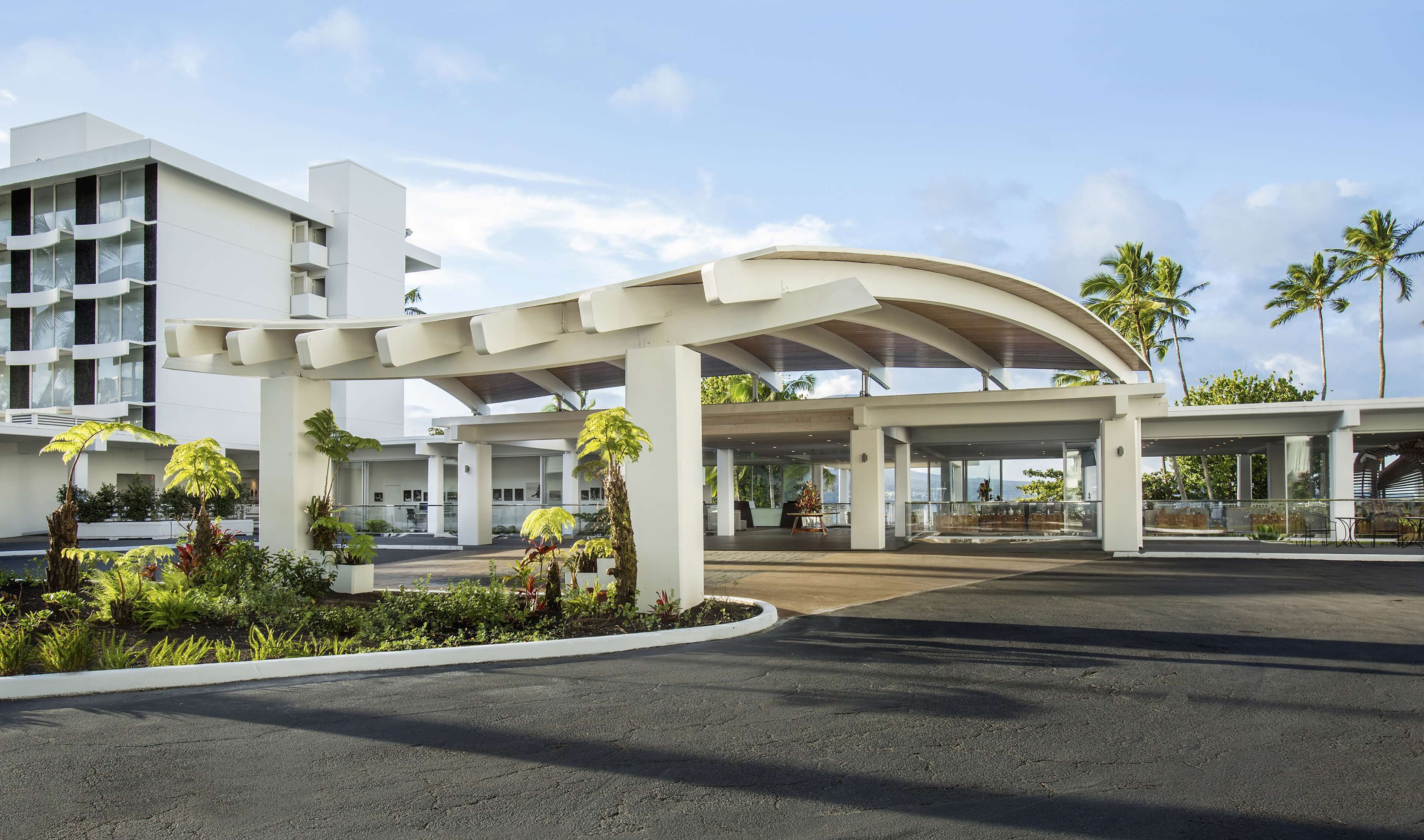 Una entrada de hotel moderna con un paseo techado, palmeras y vistas de los edificios circundantes.