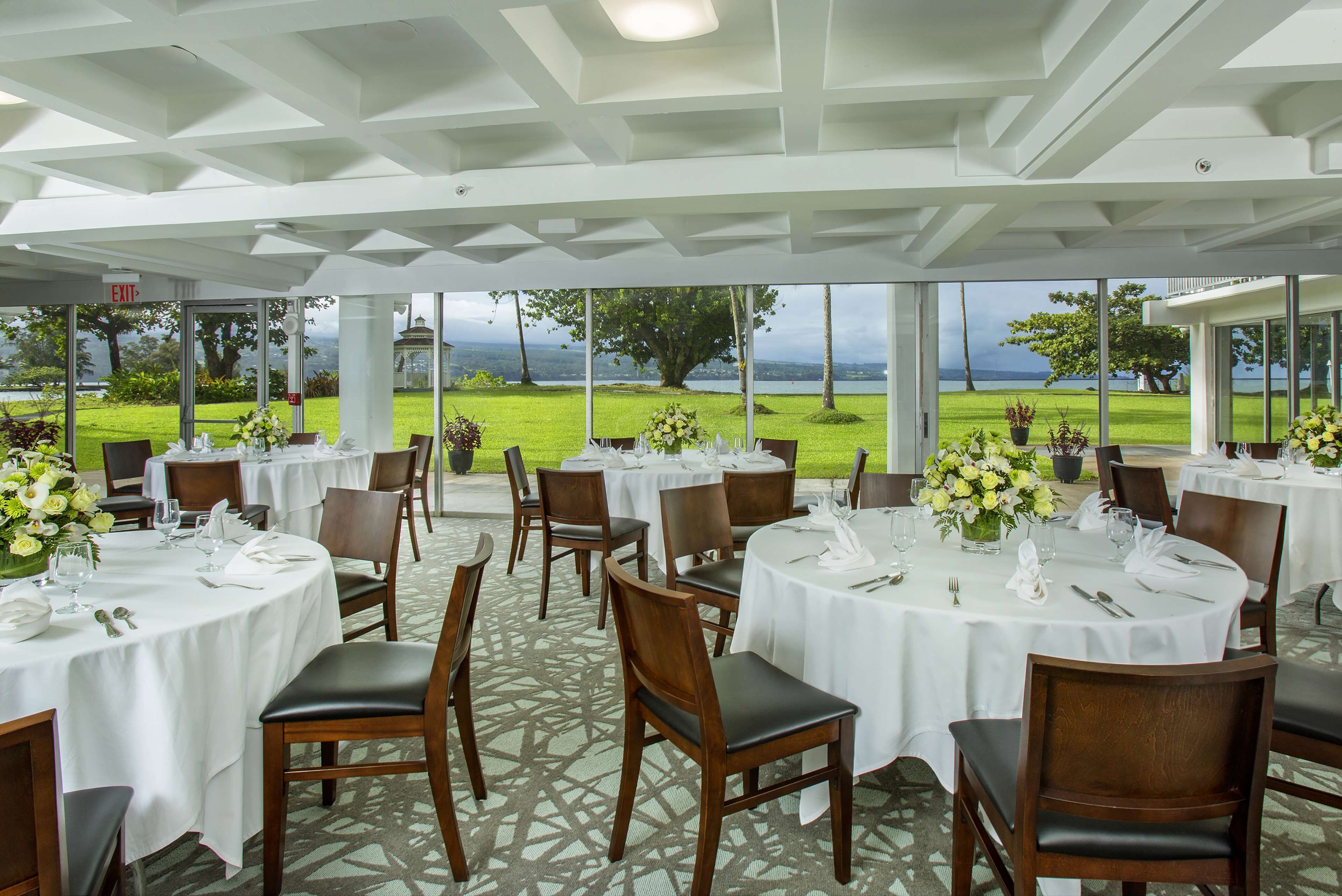 Un ristorante con tavoli rotondi, tovaglie bianche e sedie. Sui tavoli ci sono piante in vaso. Il ristorante ha ampie finestre che offrono vista su una zona verde e un corpo d'acqua.