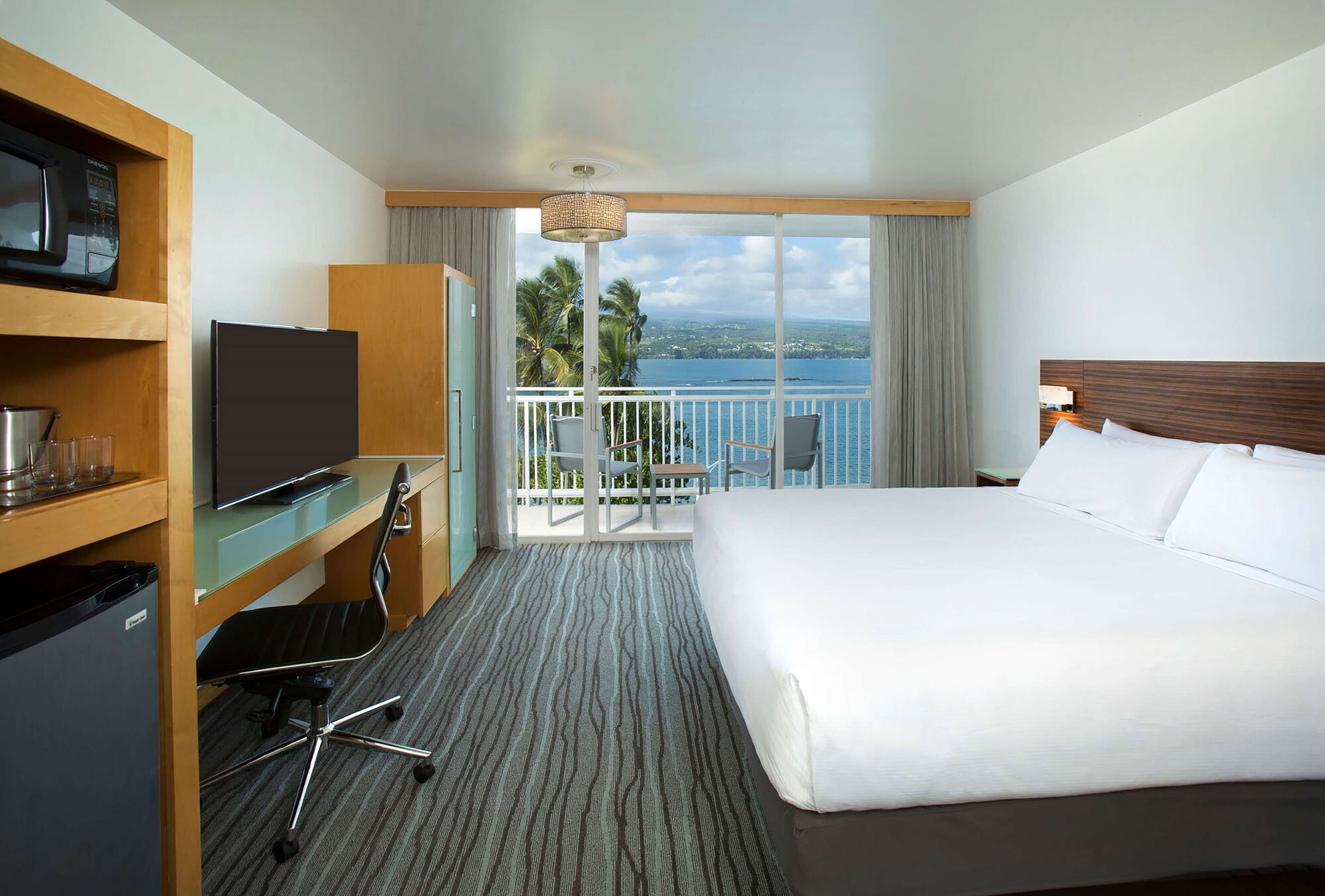 Een hotelkamer met een bed, een bureau met een televisie, een minibar en een balkon met uitzicht op zee.