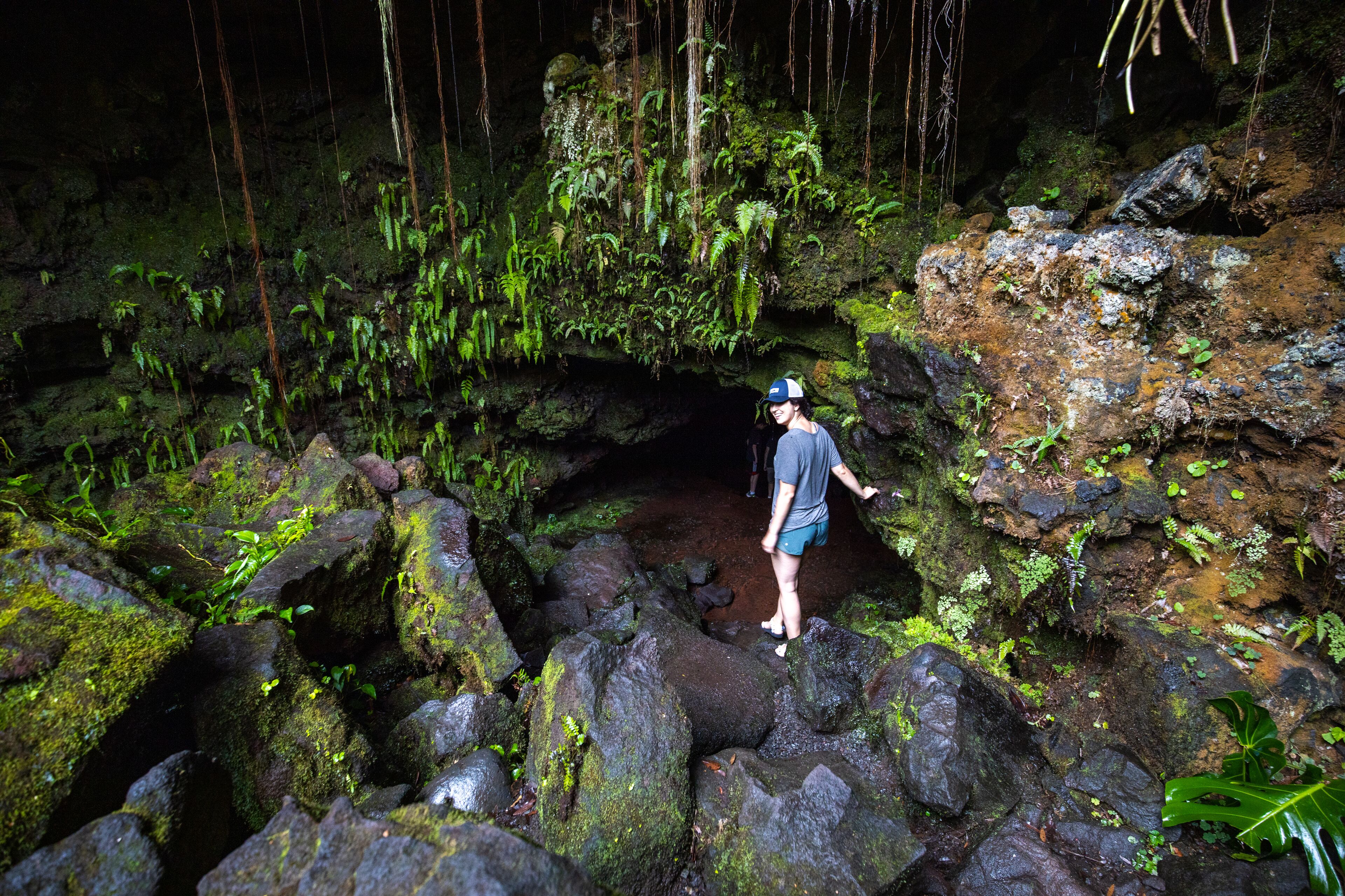 Une personne se tient dans une grotte naturelle, les rochers étant recouverts de mousse et de la végétation pendant des lianes.