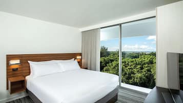 A hotel room with a large bed, a wooden headboard, a nightstand, a TV, and a view of a forest through a sliding glass door.