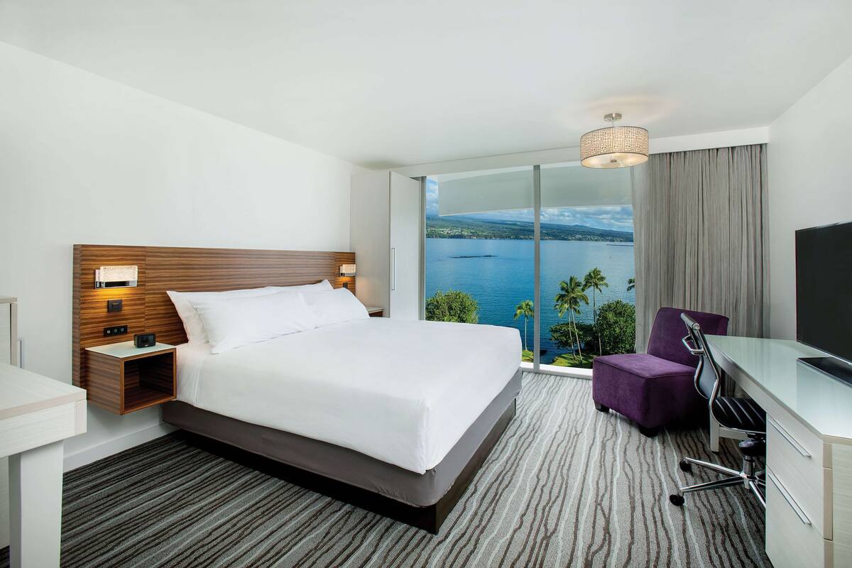 Ein Hotelzimmer mit einem großen Bett, einem Schreibtisch mit Stuhl, einem Fernseher, einem Nachttisch und Blick aufs Meer.