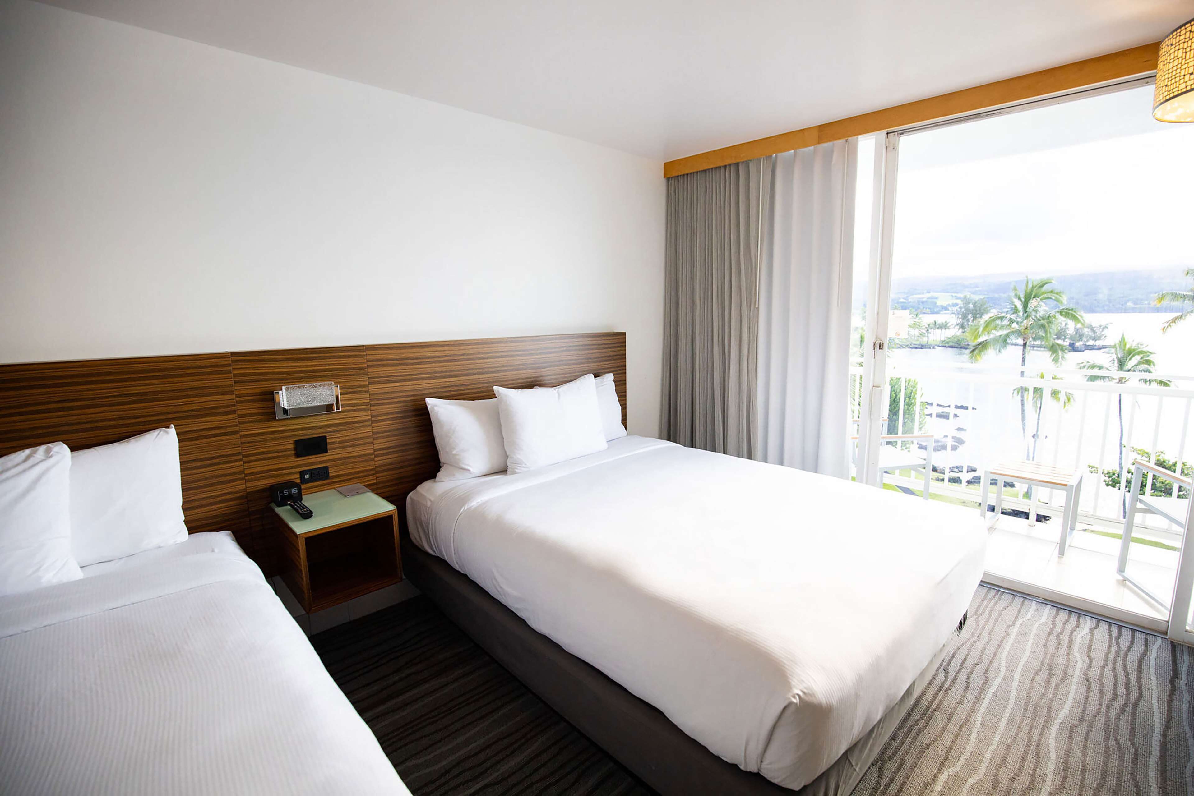 A hotel room with two beds, a wooden headboard, a nightstand with a phone, and a large window with a view of palm trees and buildings.