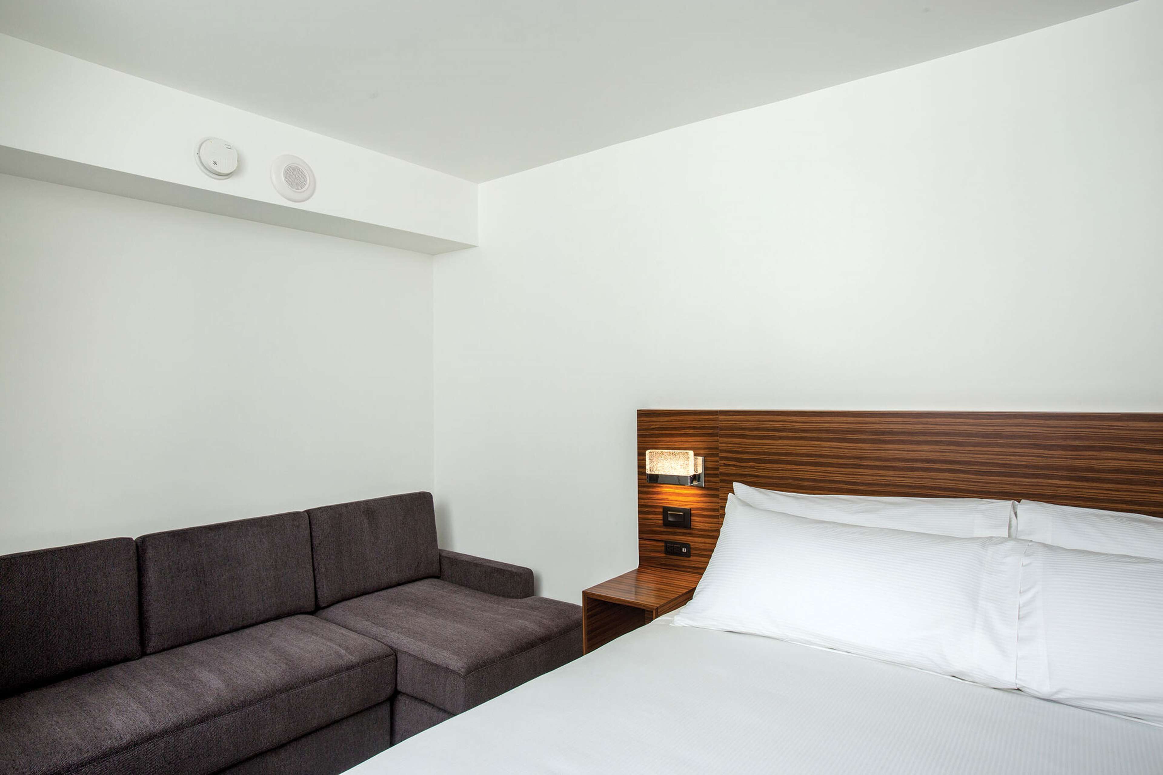 A hotel room with a bed, a wooden headboard, a nightstand, a lamp, and a gray sofa.