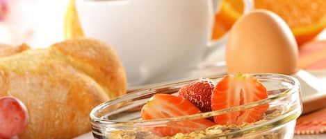 Un desayuno con un bol de granola coronado con fresas, un croissant, una taza de café y una naranja entera.