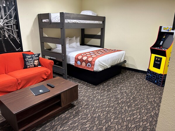 A bunk bed with a patterned bedspread, a red sofa with a pillow, a small wooden table with a remote control and a laptop, and a Pac-Man arcade machine. - Super 8 by Wyndham Gettysburg (Gettysburg)