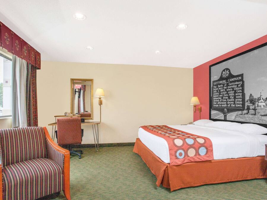 A hotel room with a bed, a chair, a desk, a lamp, and a framed historical map on the wall.