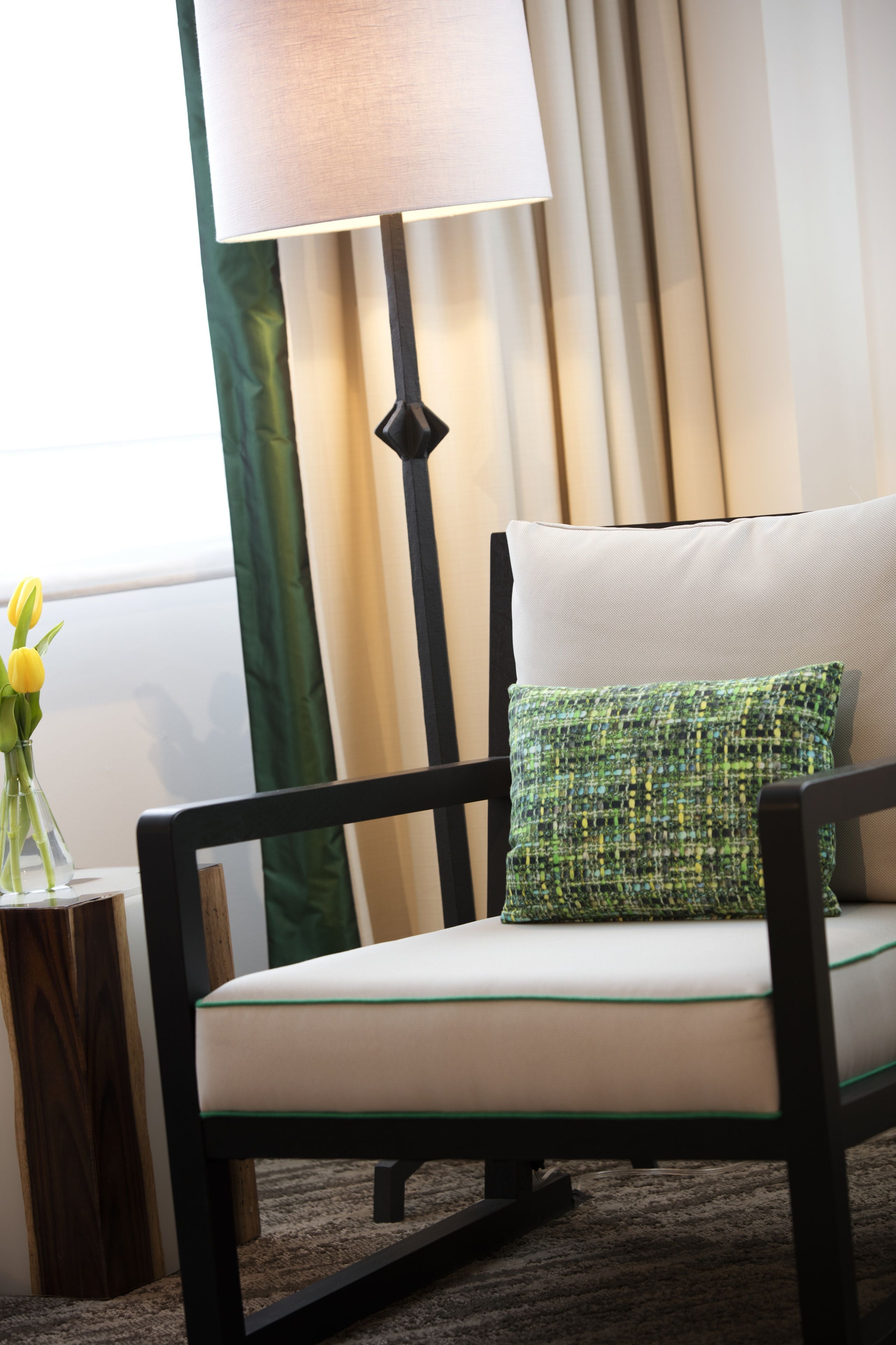 A modern armchair with a green and white patterned pillow, positioned next to a floor lamp and a vase with yellow flowers.