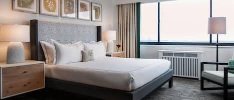 Quarto de hotel moderno com uma cama grande, uma mesa de cabeceira com luminária, uma cadeira e uma janela com vista.