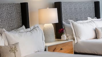 Quarto de hotel com duas camas, uma mesa de cabeceira com luminária, um vaso decorativo e cabeceira estampada.