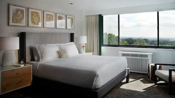 A modern hotel room with a large bed, a nightstand, a lamp, a view of the city through large windows, and framed artwork on the wall.