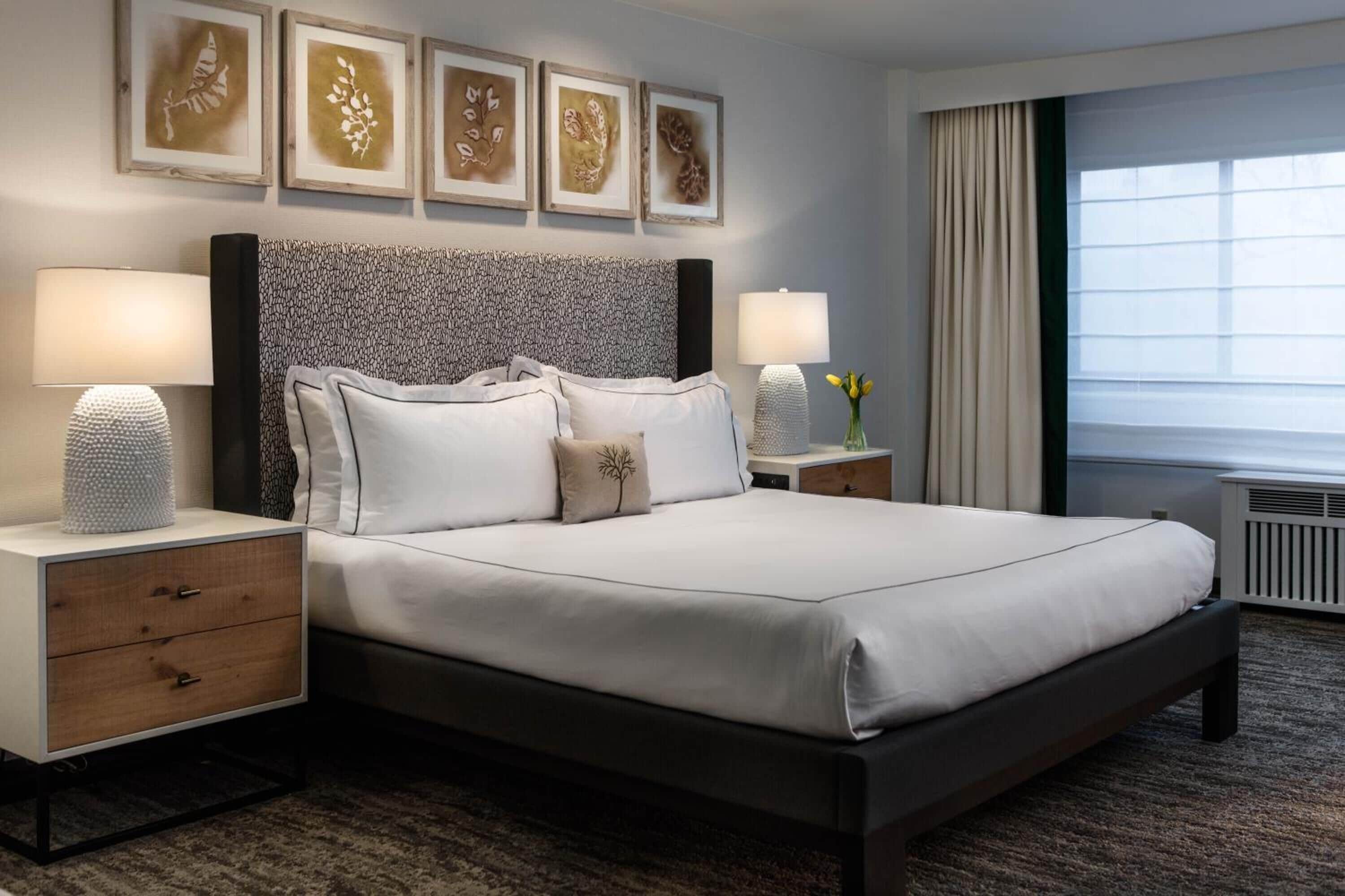 A hotel room with a large bed, two bedside tables, a headboard with a patterned design, and a window with curtains.