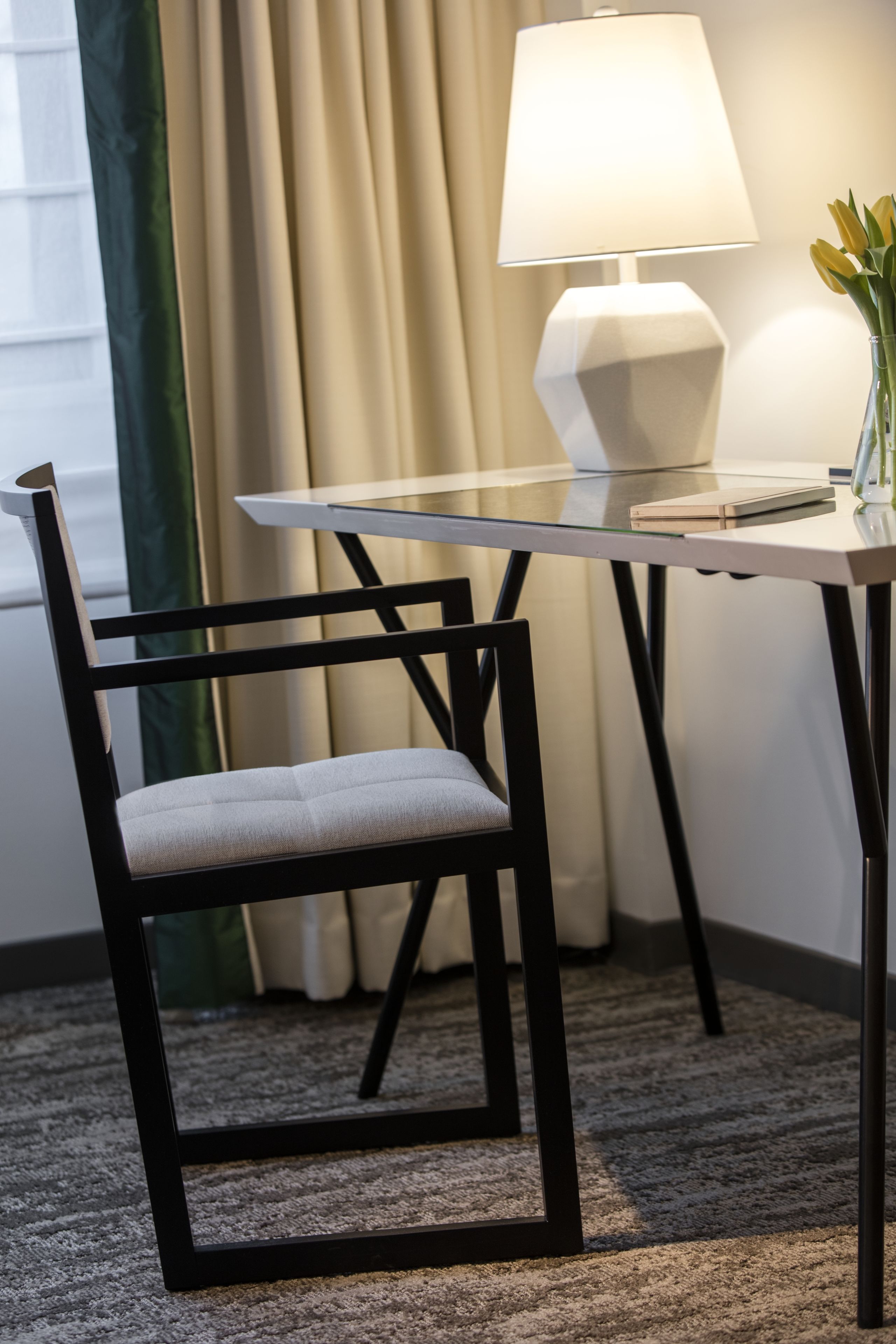 Una habitación de hotel moderna con una mesa de vidrio, una silla negra, una lámpara blanca y un florero con flores amarillas.