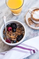 A bowl of granola with berries and a glass of orange juice on a marble surface.