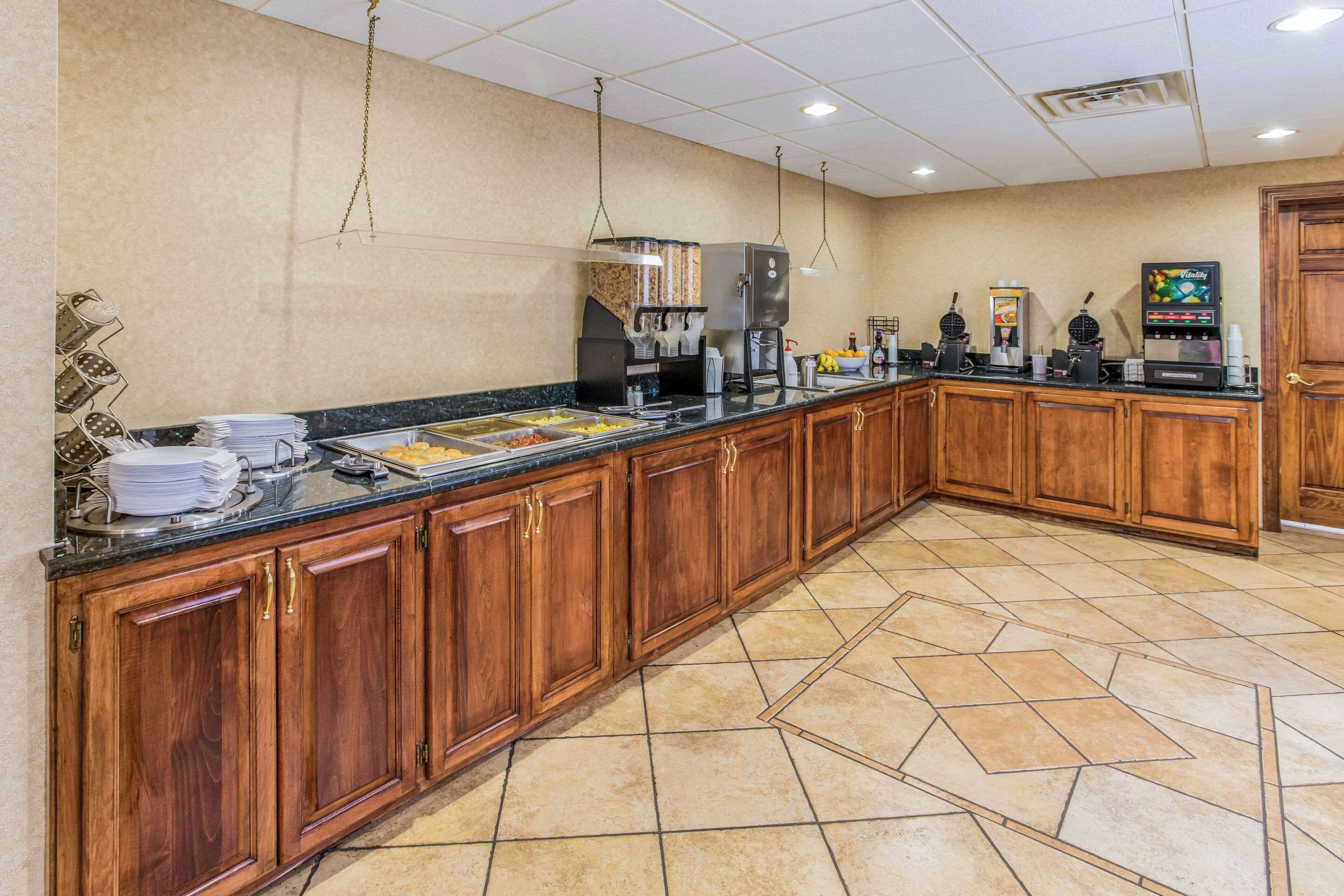 Una cocina de hotel con armarios de madera, encimera de mármol y diversos electrodomésticos, como cafeteras y dispensador de alimentos.
