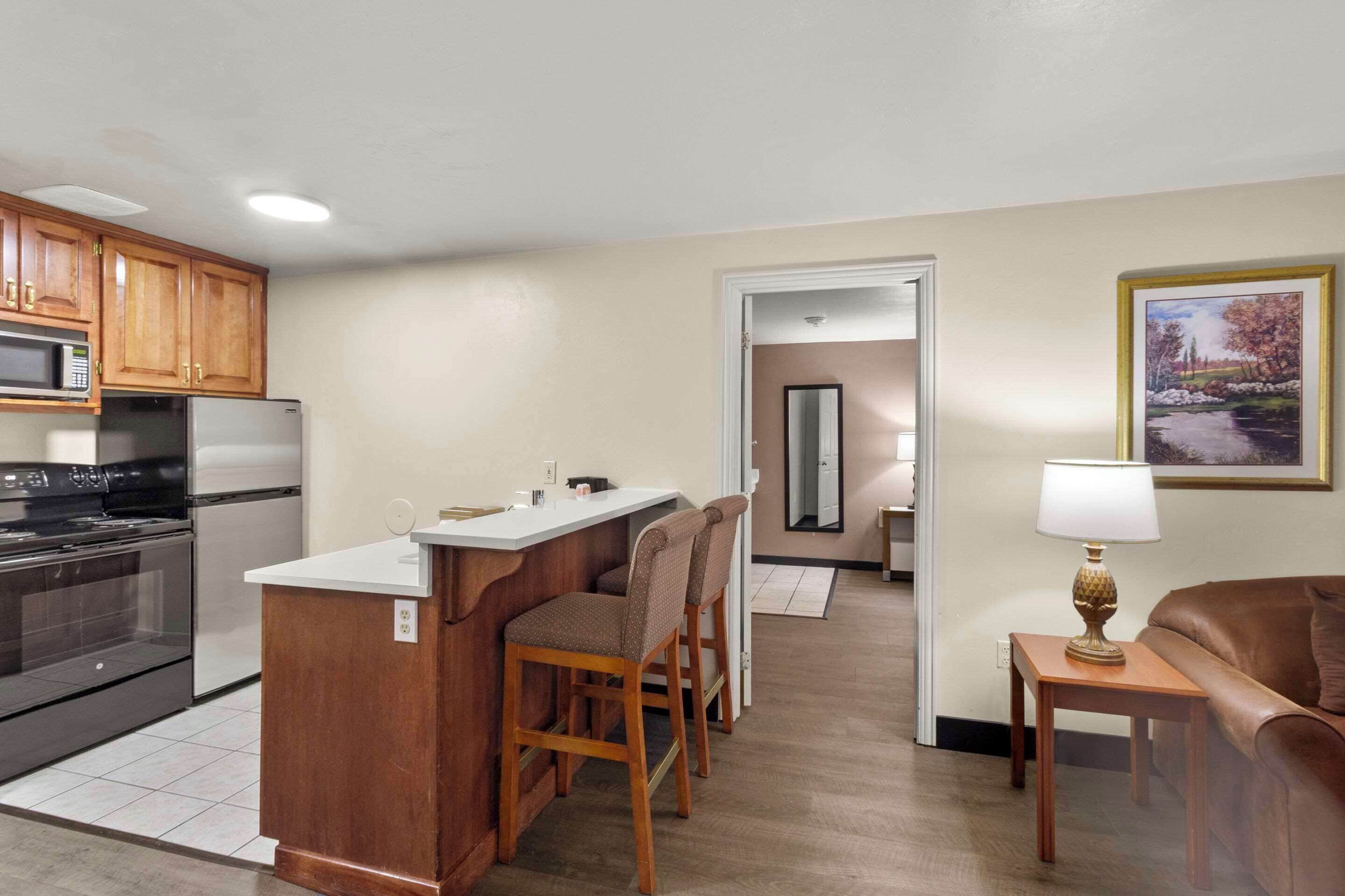 A compact kitchen with wooden cabinets, a built-in microwave, and a refrigerator. There is a small island with bar stools, a dining table, and a lamp. A brown leather sofa is visible in the living area.