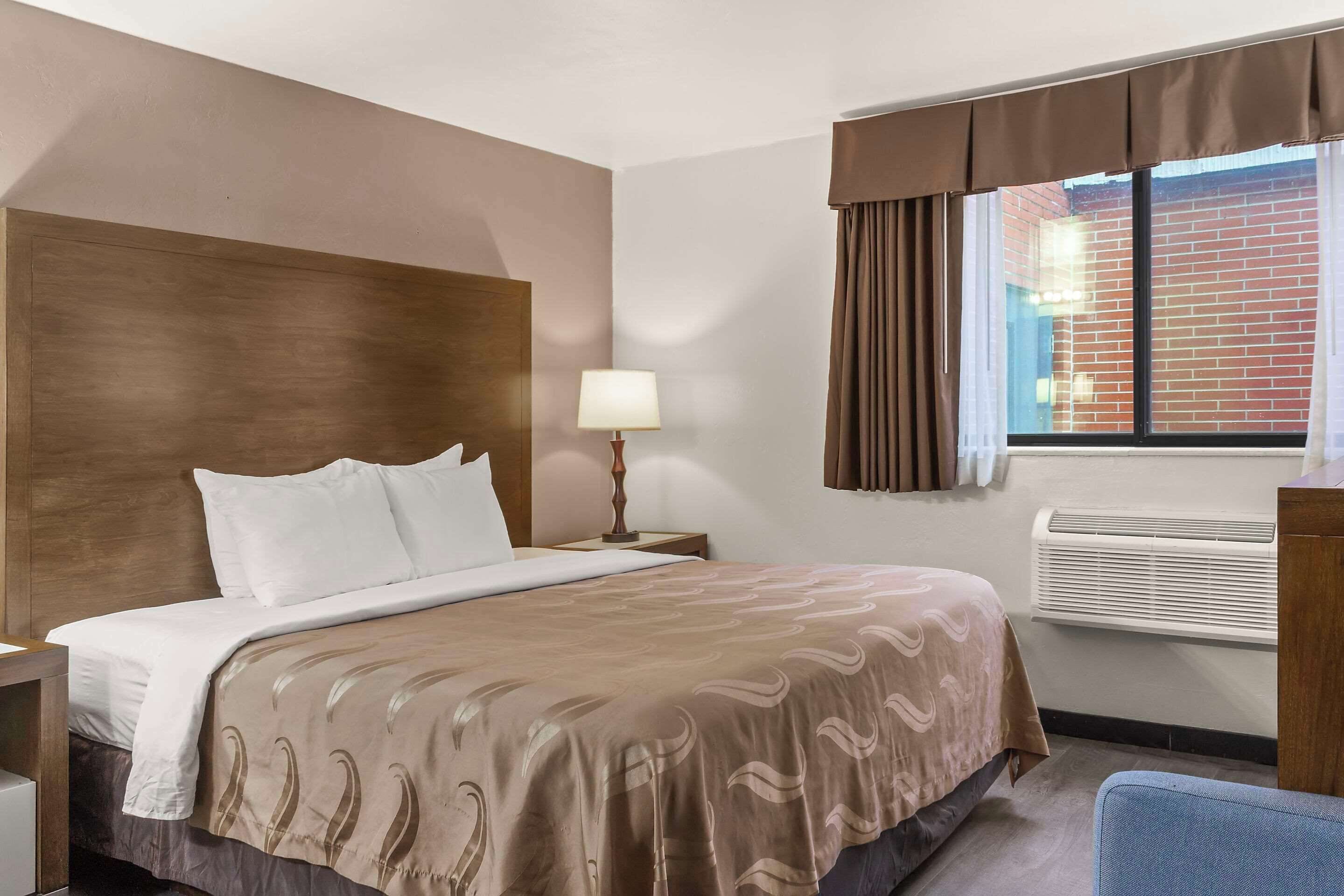 A hotel room with a large bed, a bedside table with a lamp, an air conditioning unit, and a view of a brick building through a window with curtains.