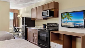 A hotel room with a kitchenette including a microwave, stove, refrigerator, and a flat-screen TV mounted on the wall.
