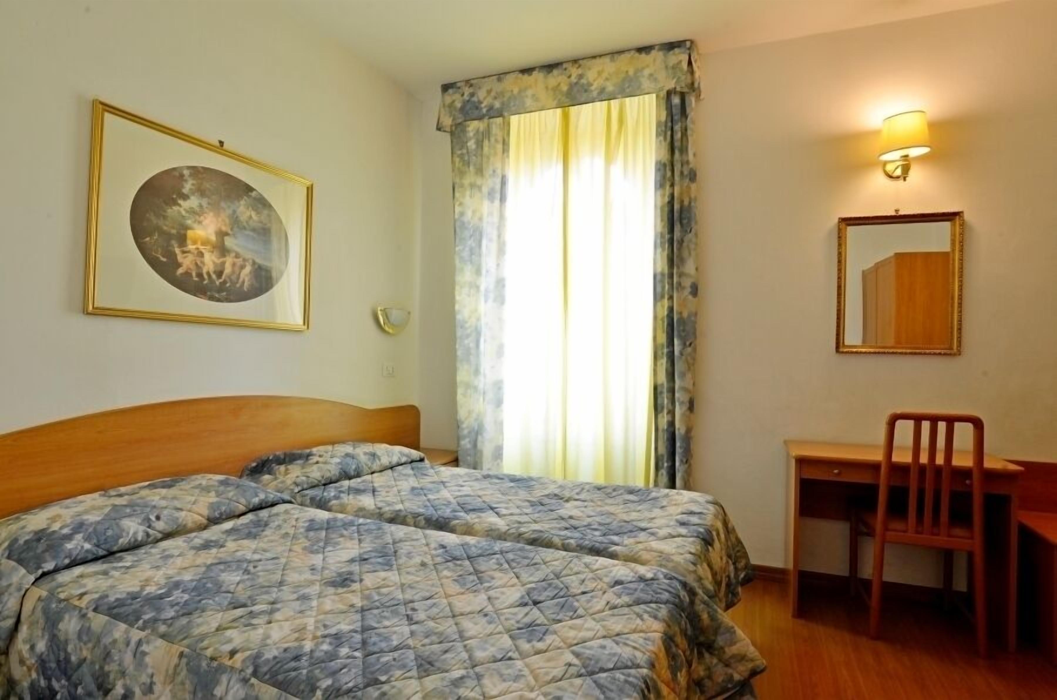 A room with two beds, a desk, a chair, a wall-mounted lamp, and a framed picture.
