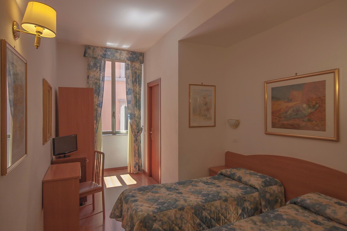 A hotel room with a bed, a desk with a television, a chair, and a window with curtains.