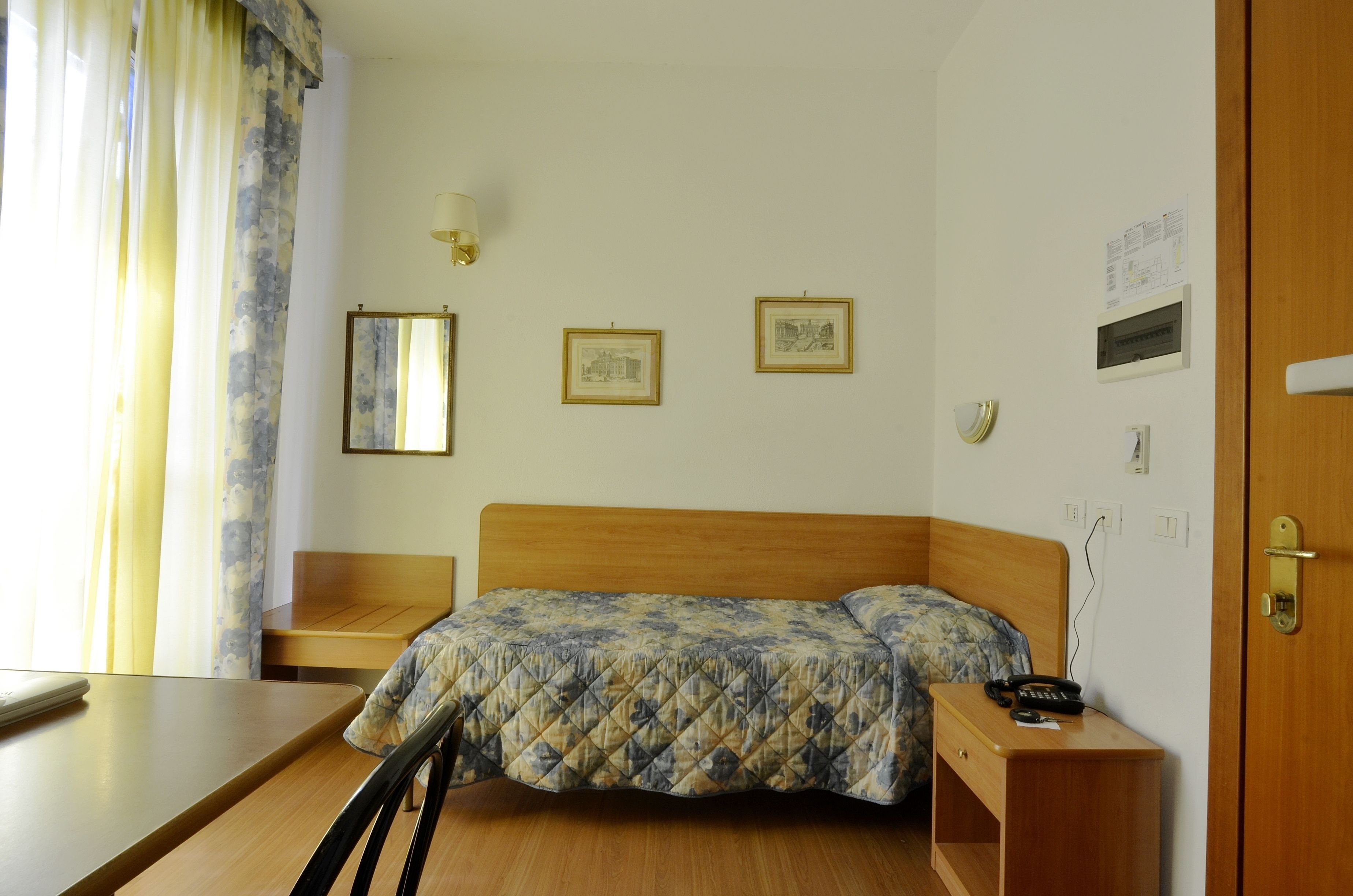 Une chambre d’hôtel avec un lit en bois, une table de chevet avec une lampe et une chaise. Des tableaux encadrés ornent le mur, et il y a une fenêtre avec des rideaux.