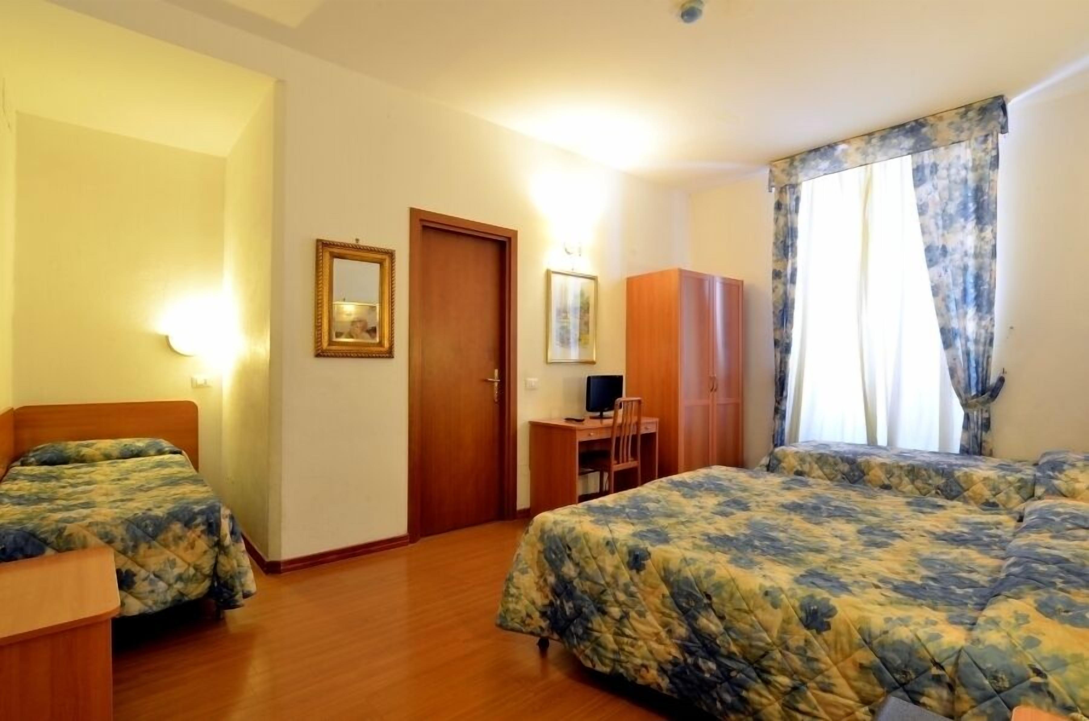 A hotel room with two beds, a desk with a computer, a wardrobe, and a window with curtains.