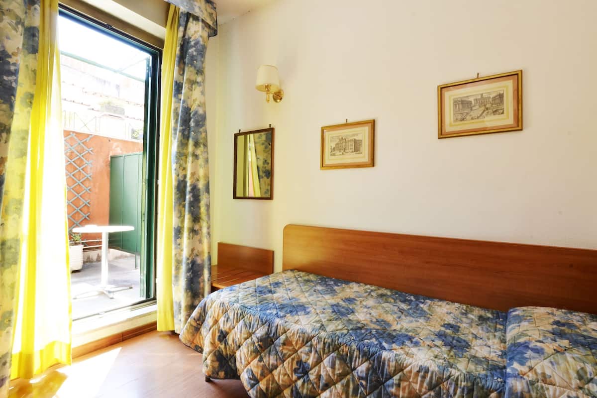 A hotel room with a bed, a chair, a table, and a window with curtains. There are two framed pictures on the wall.