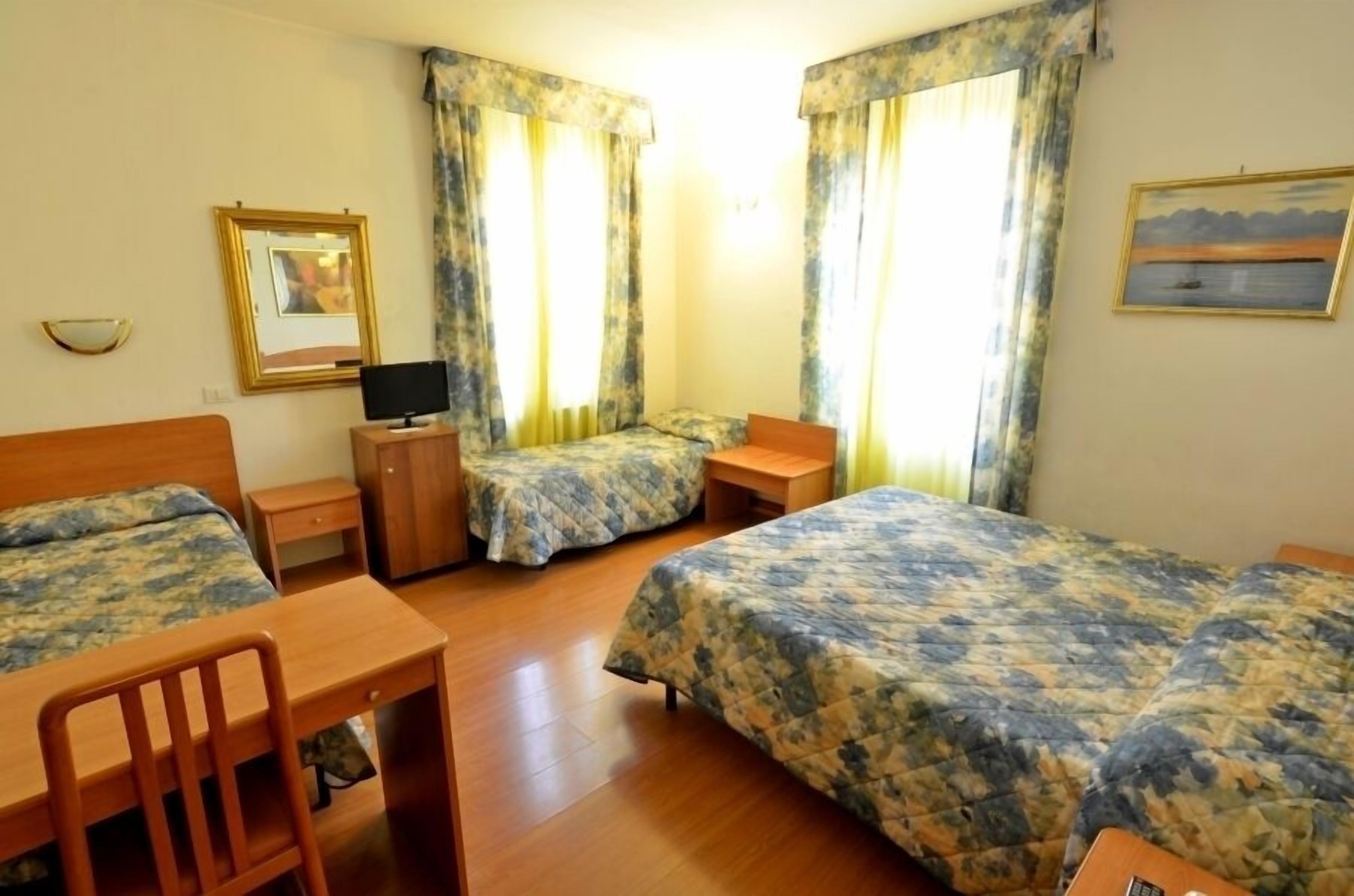 Hotellihuone, jossa on kaksi erillistä sänkyä, puinen työpöytä, tuoli, pieni pöytä, televisio ja verhot.