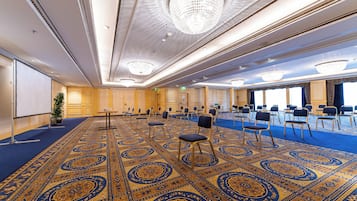 Sala de conferências com carpete estampado, fileiras de cadeiras e tela de projeção.