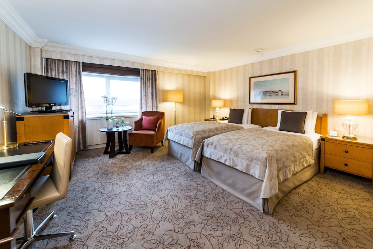 Ein Hotelzimmer mit zwei Betten, einem Schreibtisch mit Fernseher, einem Sessel, zwei Nachttischen mit Lampen und einem gerahmten Bild an der Wand.