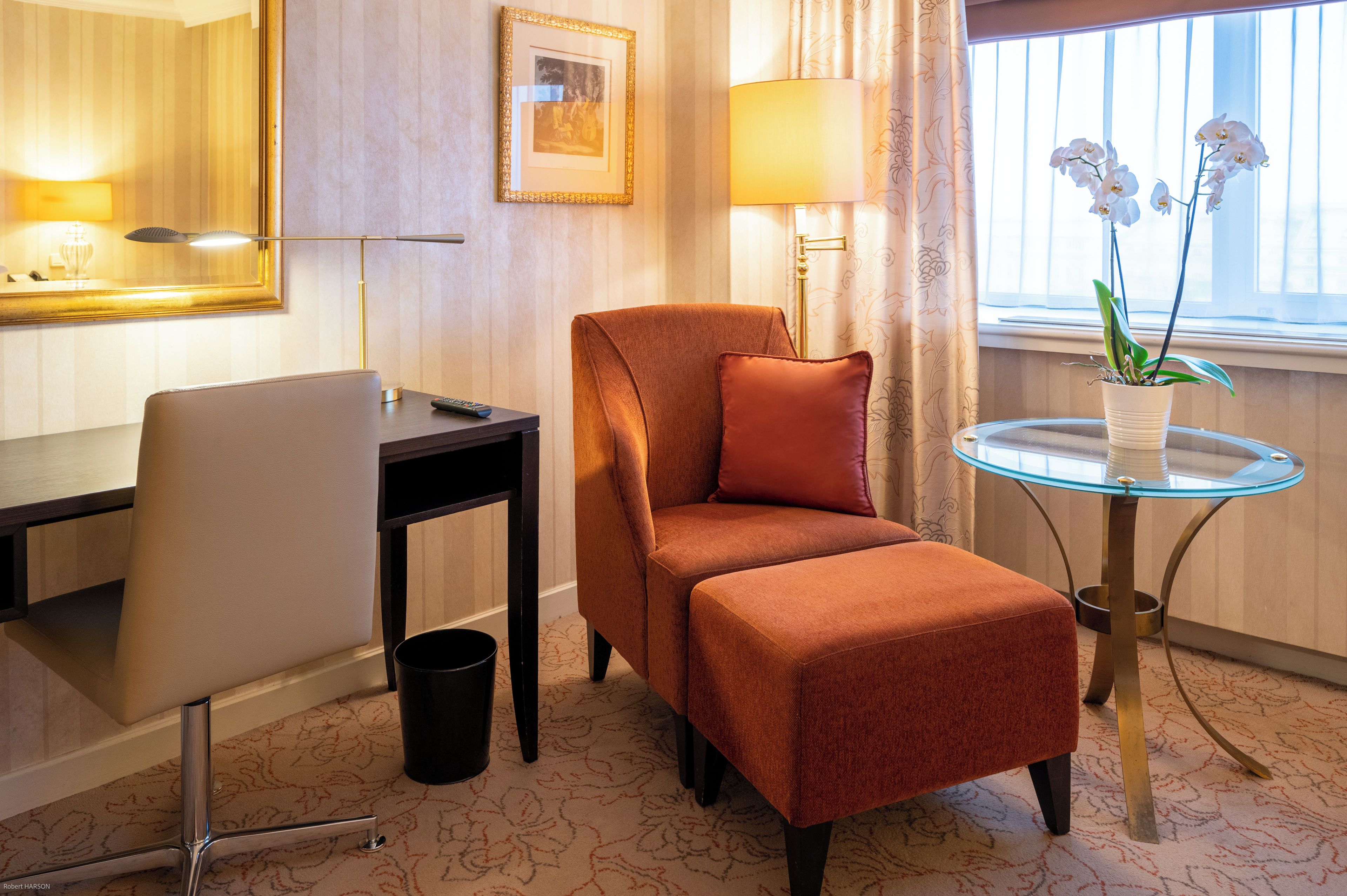 Ein Hotelzimmer mit einer Couch, einem kleinen Tisch mit Glaseplatte, einer Lampe, einem Bilderrahmen und einem Fenster mit Vorhängen.