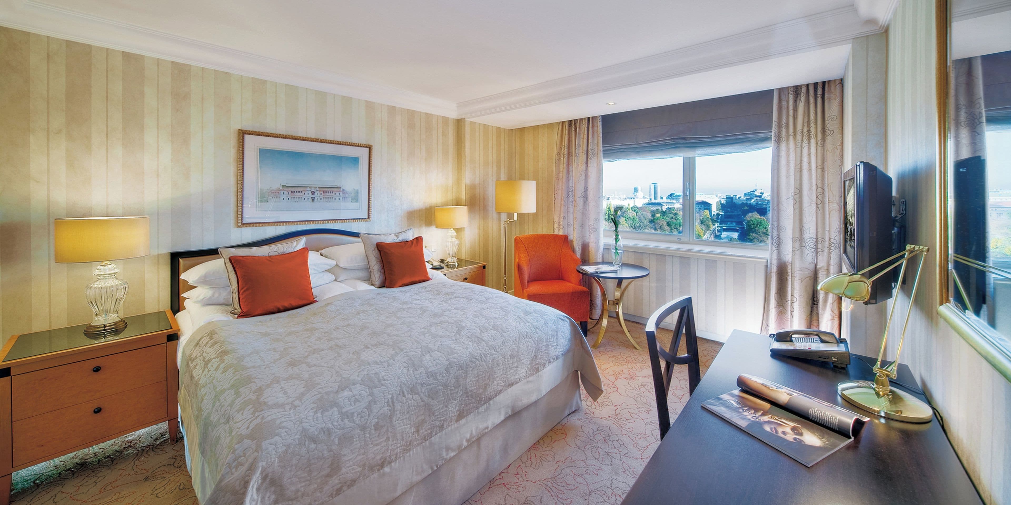 A hotel room with a large bed, two bedside tables, a desk with a chair, a television, and a window with a city view.