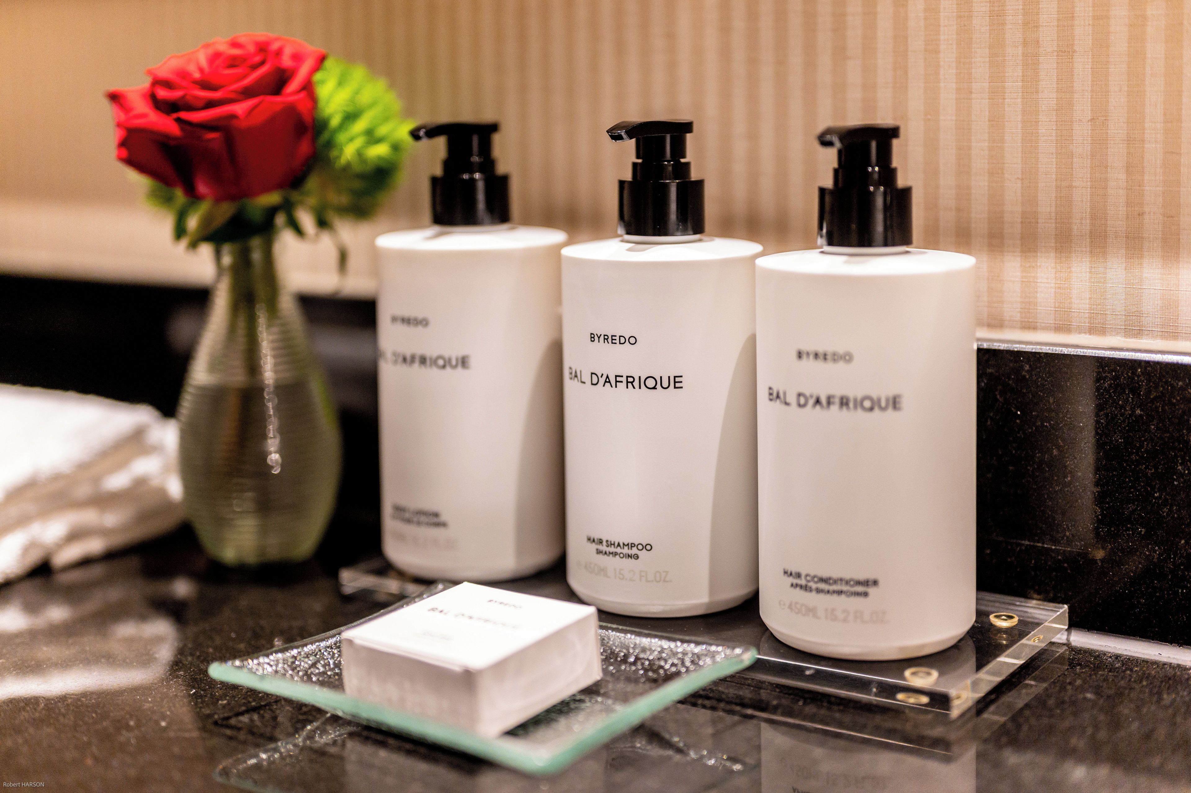 Três frascos brancos com bombas pretas rotulados 'BYREDO BAL D'AFRIQUE' sobre uma bancada. Uma rosa vermelha e uma pequena caixa branca também estão visíveis.