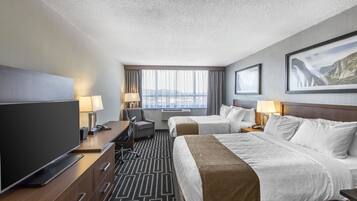 A hotel room with two beds, a TV, a desk with a lamp, and a view of the mountains through the window.