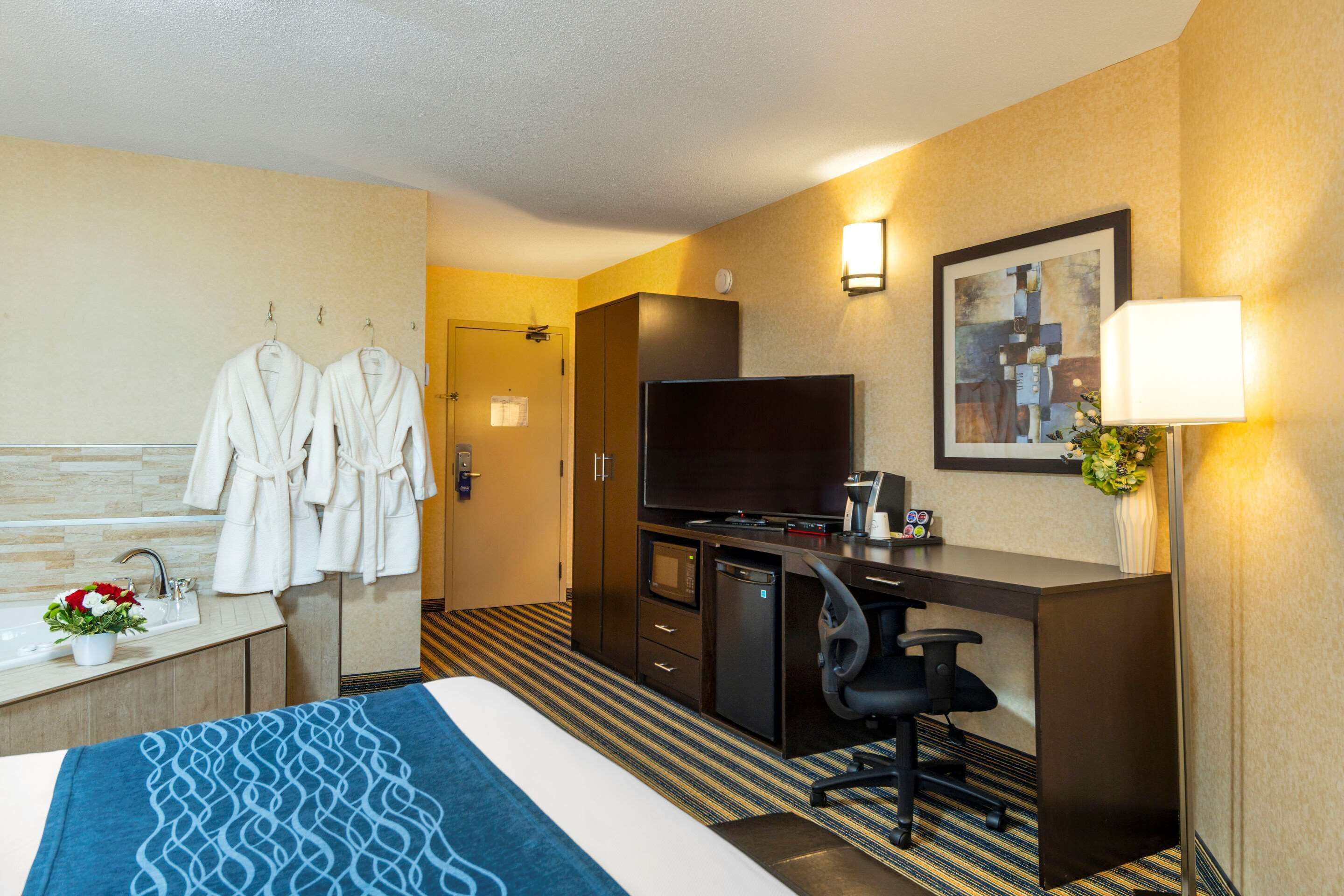 A hotel room with a bed, a desk, a TV, a minibar, a bathroom with a sink and towels, and a painting on the wall.
