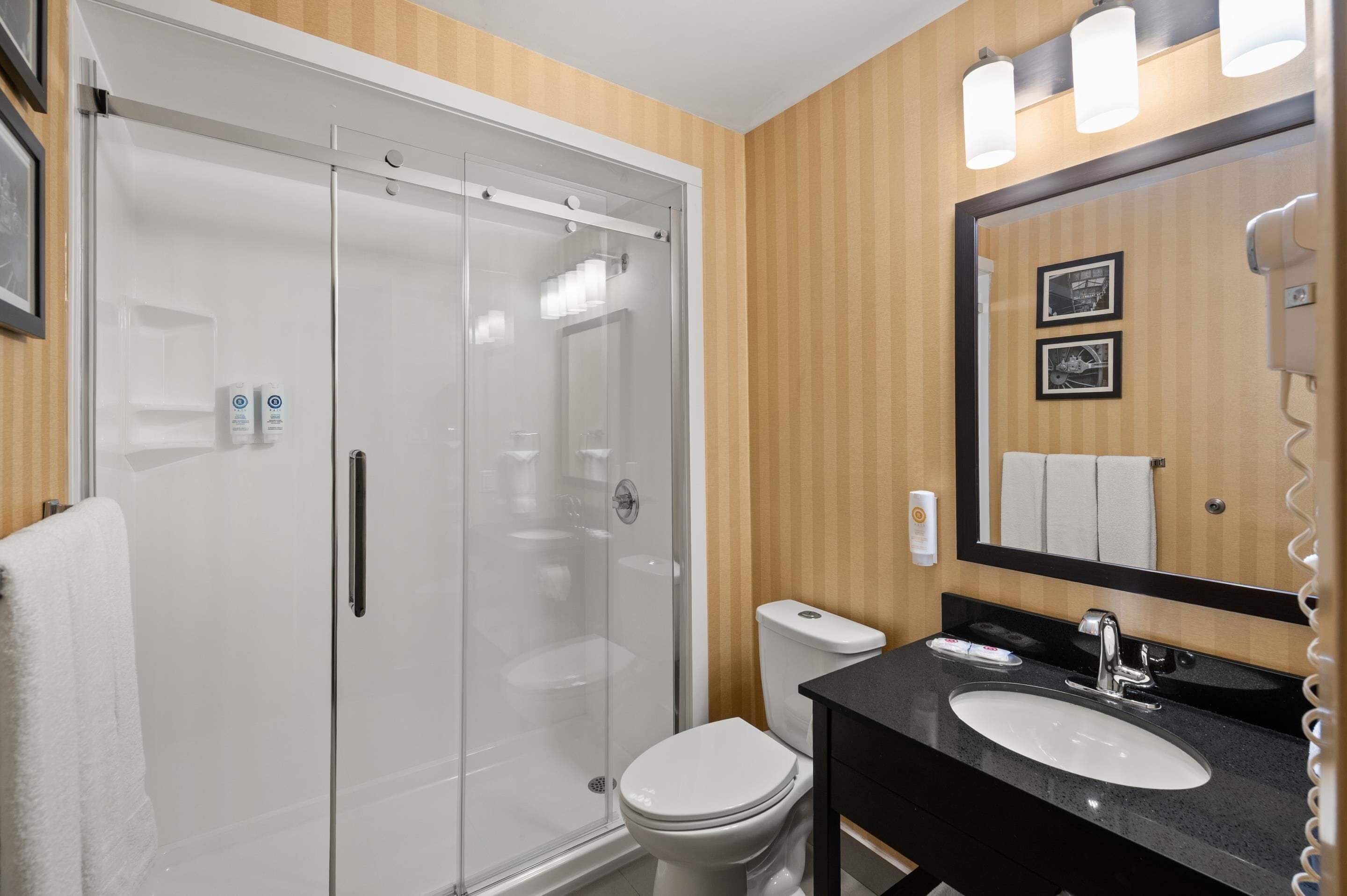 A modern bathroom with a glass-enclosed shower, a white toilet, a black sink, and a mirror with lights.