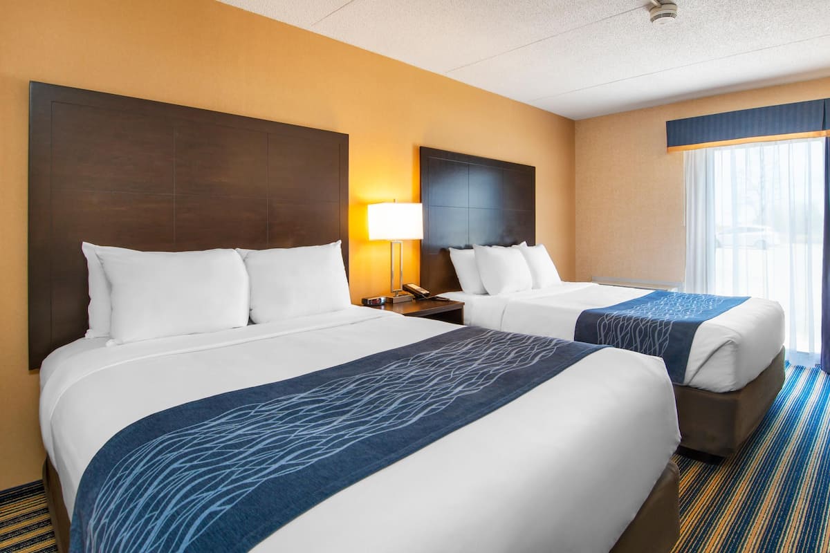 A hotel room with two beds, a nightstand with a lamp, and a large window with curtains.