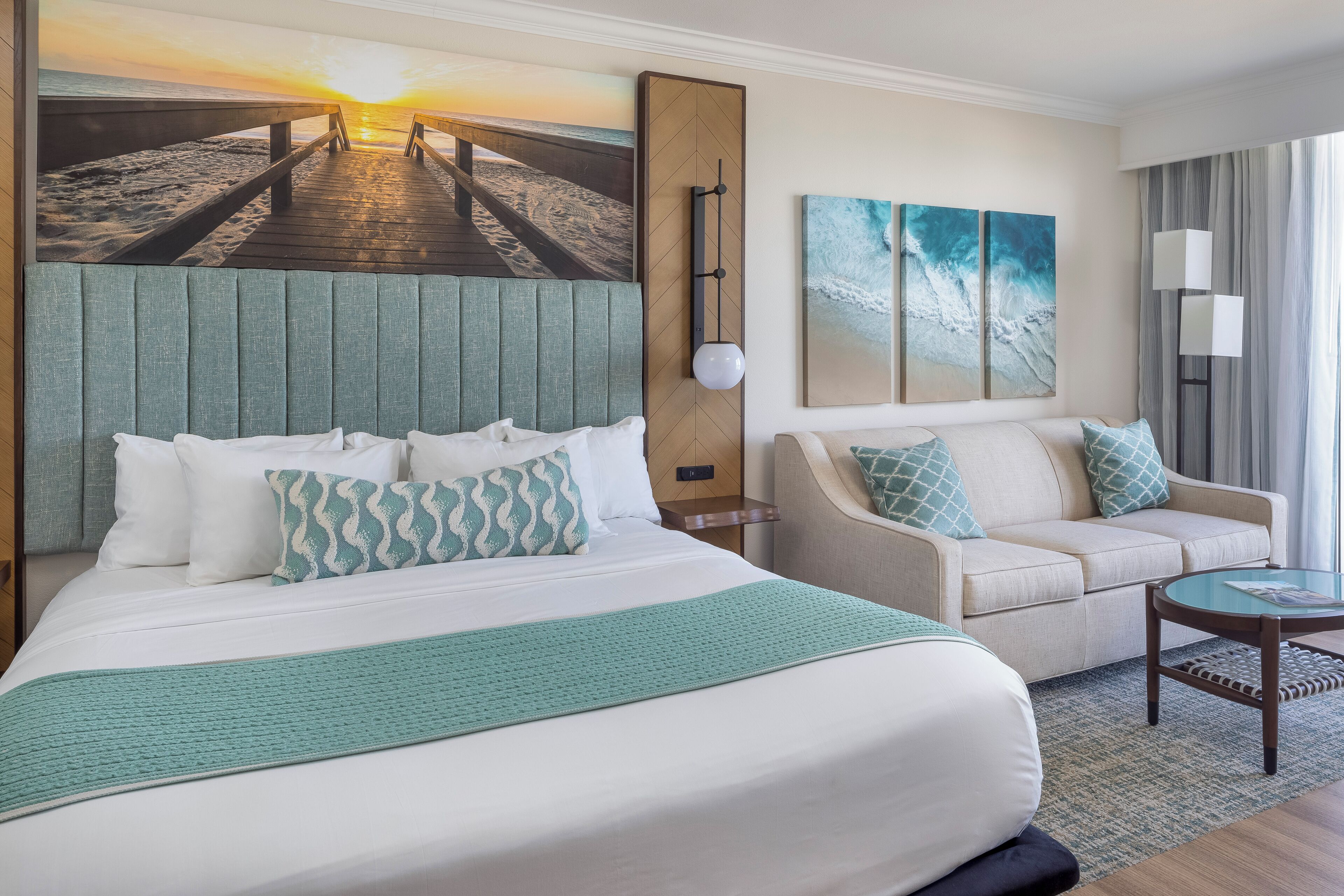 Camera d'albergo con un letto, un divano, un tavolino da caffè e opere d'arte raffiguranti una spiaggia.