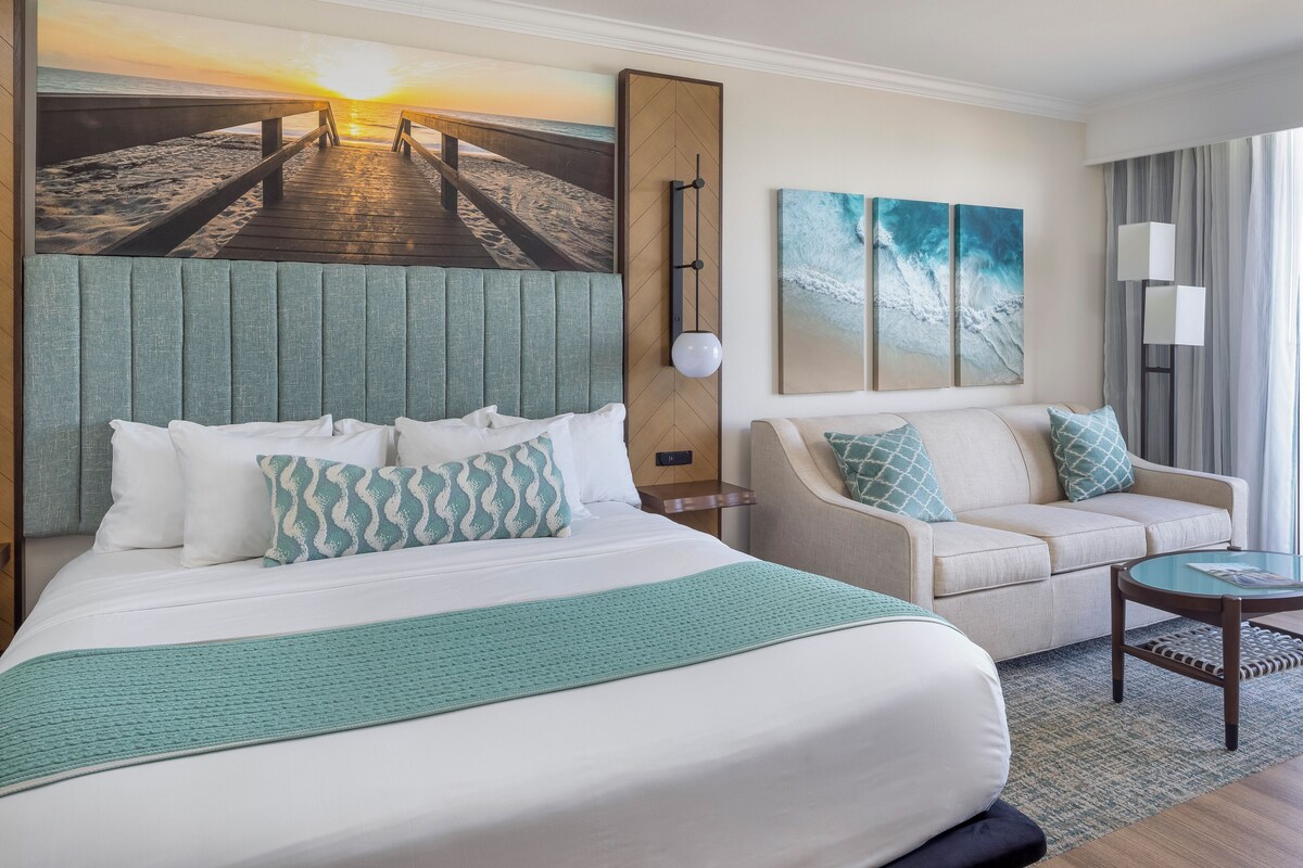 A hotel room with a bed, a sofa, a coffee table, and artwork depicting a beach scene.
