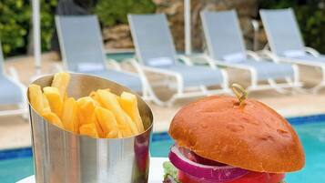 Un pasto con hamburger e patatine fritte vicino a una piscina, sullo sfondo lettini e ombrelloni.