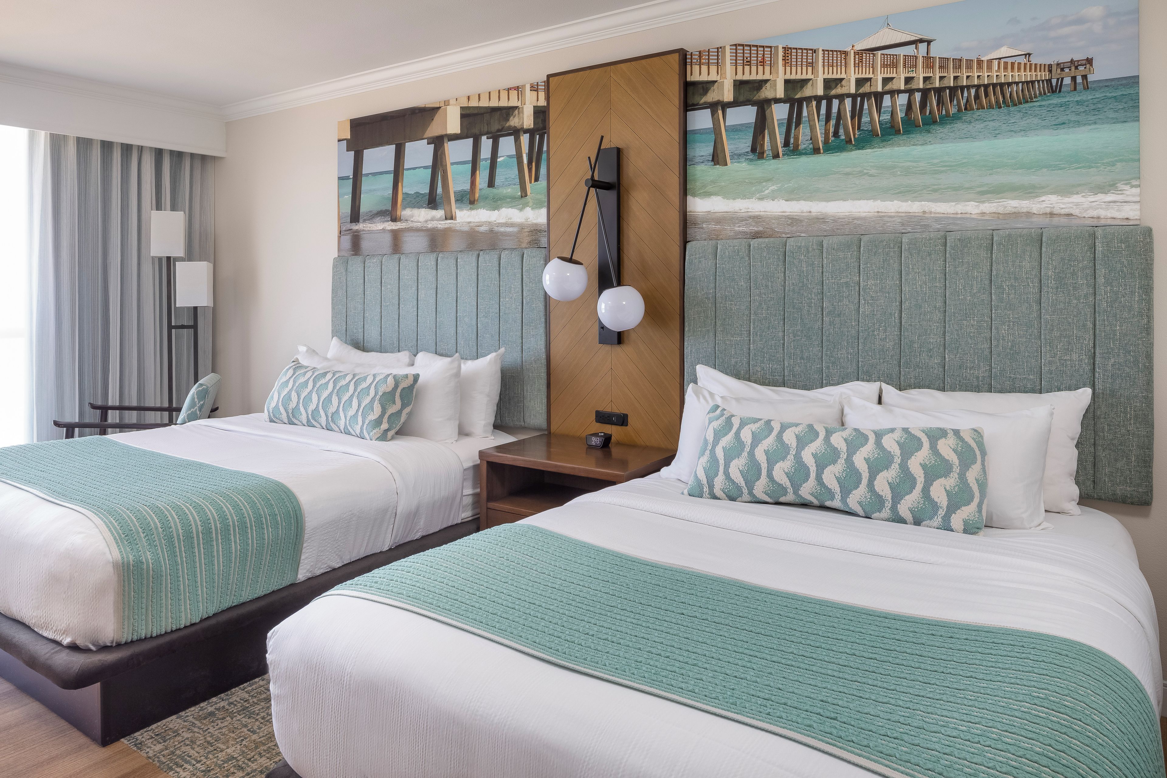 A hotel room with two beds, a large window with curtains, a painting of a pier, and a nightstand with a lamp.