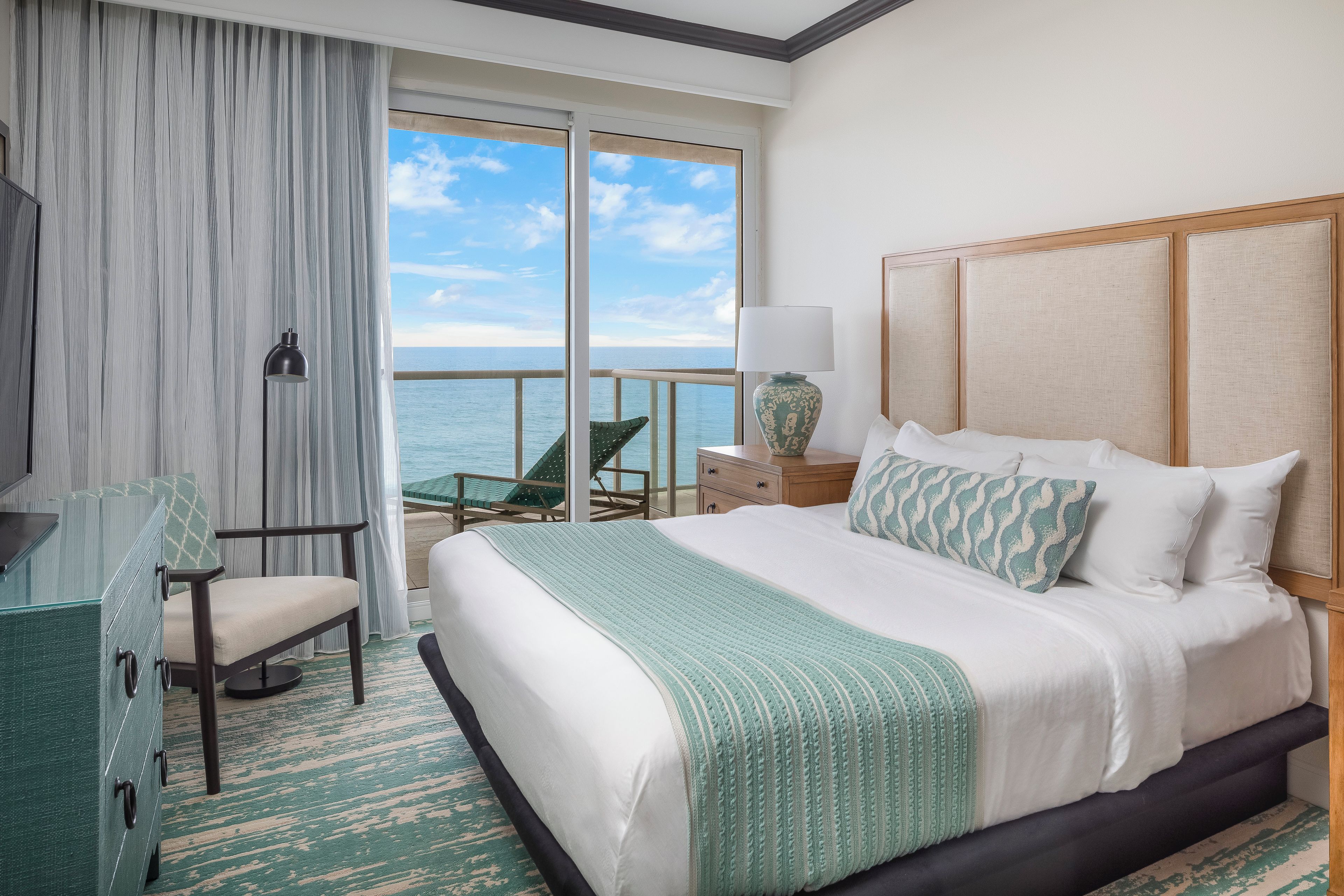 A hotel room with a large bed, a chair, a desk, and a view of the sea.