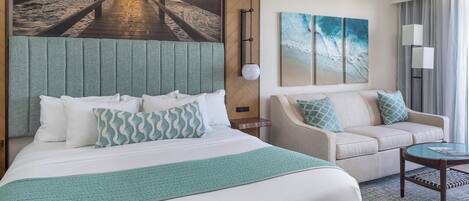 Quarto de hotel com cama, sofá, mesa de centro e obra de arte retratando uma cena de praia.