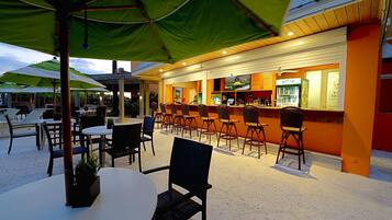 An outdoor bar area with multiple tables and chairs, green umbrellas, and a bar counter with a beer tap.