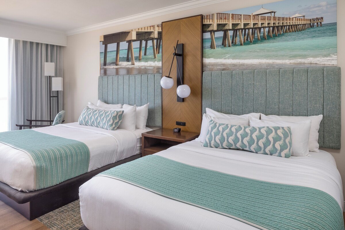 A hotel room with two beds, a large window with curtains, a painting of a pier, and a nightstand with a lamp.