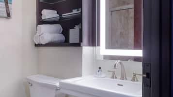 A bathroom with a toilet, sink, and a mirror with a light. There is a shelf with towels and toiletries.