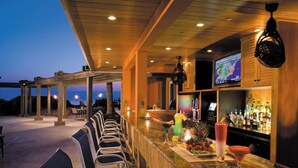 A bar area with seating and a counter, featuring a television screen and various bottles. - The Lucie (Jensen Beach)