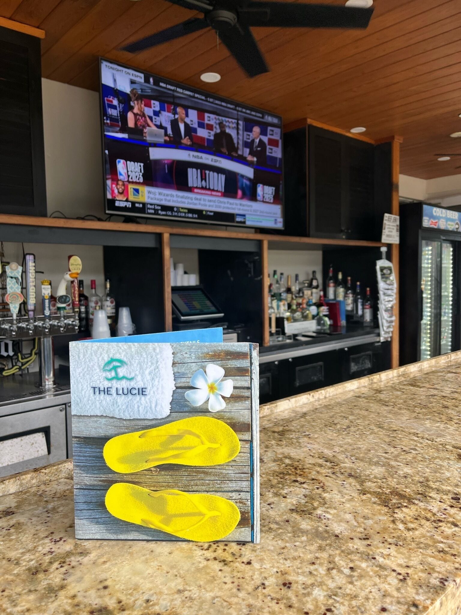 A bar area with a television displaying sports news, a counter with a menu board, and a decorative sign with a flower and slippers.