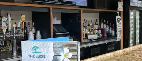 A bar area with a television displaying sports news, a counter with a menu board, and a decorative sign with a flower and slippers.