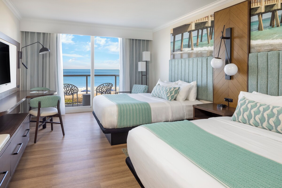 A hotel room with two beds, a desk, a chair, and a view of the ocean.